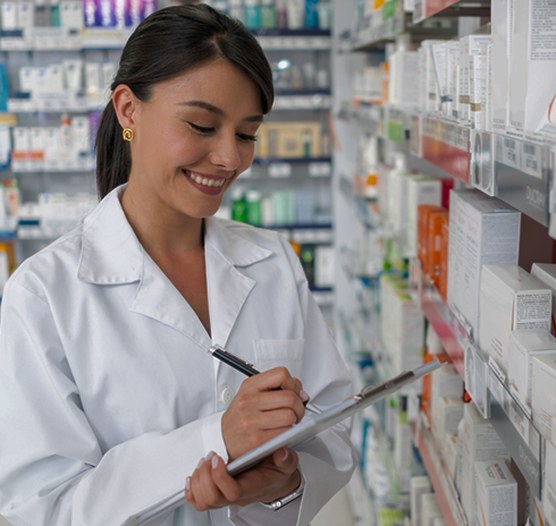 Farmacéutica sonriente con bata blanca que anota en una tabla frente a los estantes