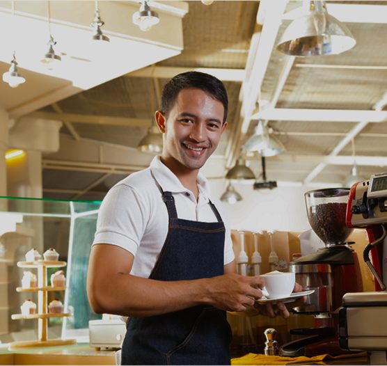 Vendedor sonriente con delantal que sostiene una taza junto a una cafetera