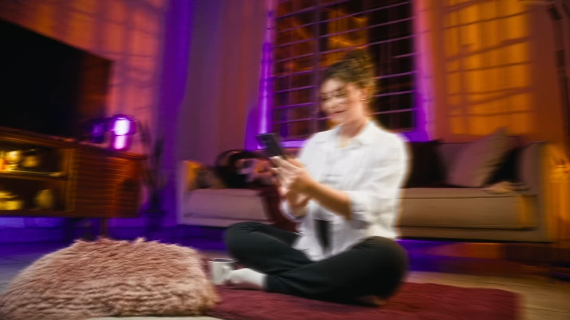 Mujer sentada en el piso de una sala iluminada en morado, mirando su teléfono