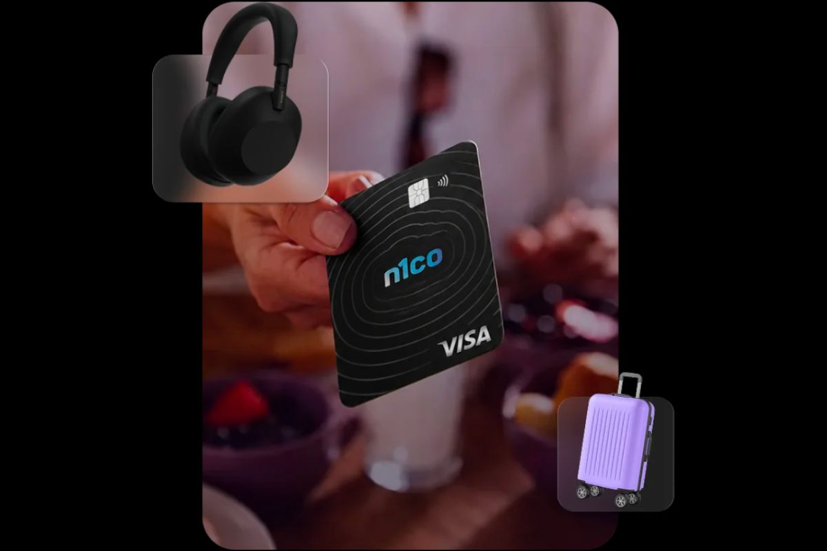 Mano que muestra una tarjeta n1co Visa, con unos audífonos y una maleta morada alrededor