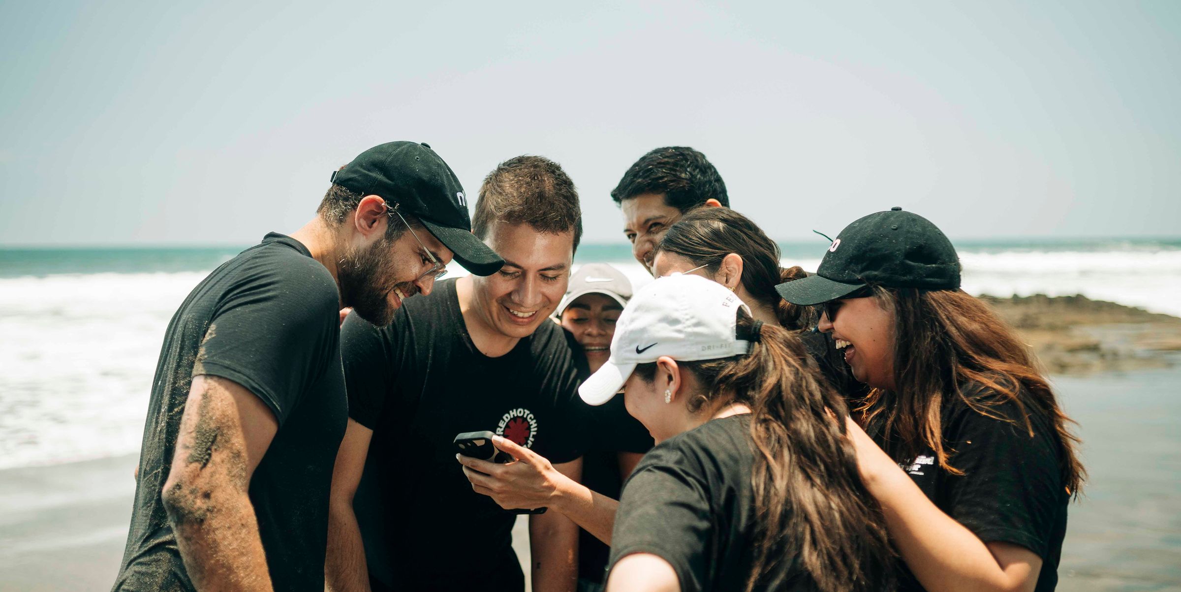 Jóvenes de Fundación n1co con gorras y camisetas negras que miran un teléfono y se ríen en la playa