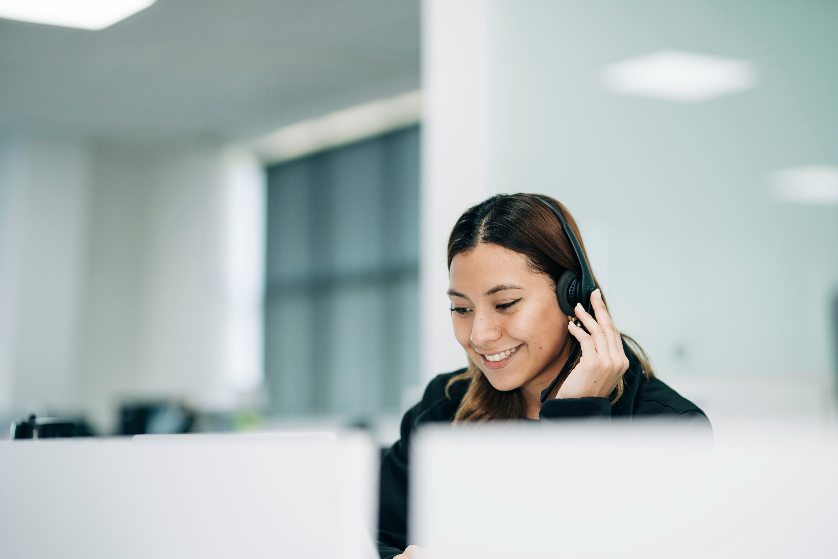 Mujer sonriente con audífonos de soporte frente a una computadora, en una oficina