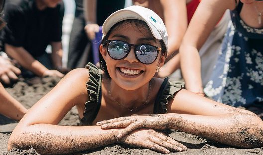 Una joven con gorra y lentes de sol sonríe recostada en la arena