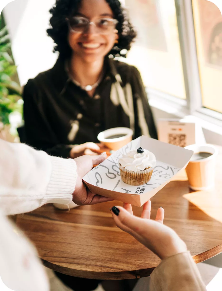 Mesera que entrega un cupcake en la mesa a una clienta sonriente, con el QR de n1co al fondo