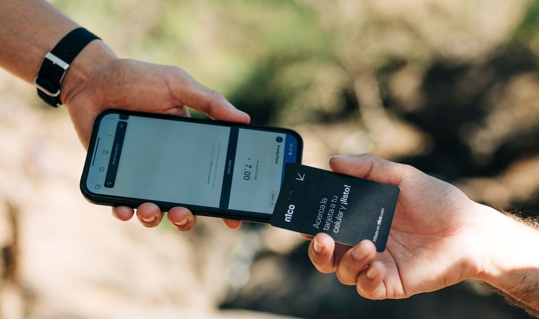 Una mano acerca una tarjeta n1co al teléfono que sostiene otra persona, al aire libre