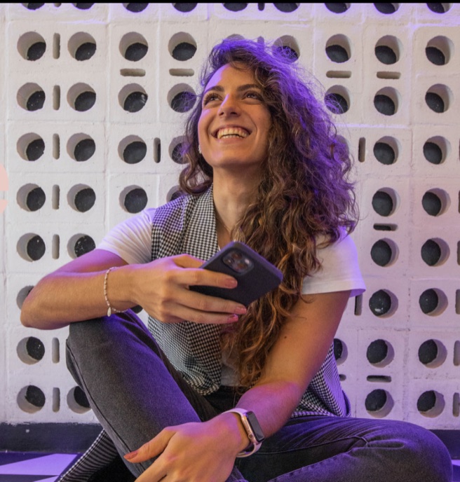 Mujer sonriente sentada frente a una pared de bloques calados, con su teléfono en la mano