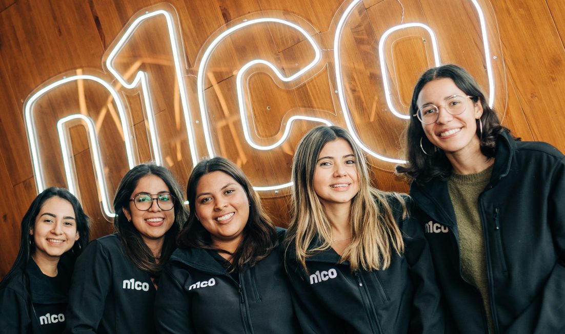 Cinco mujeres sonrientes con chaquetas de n1co frente al logo de n1co hecho con luces de neón