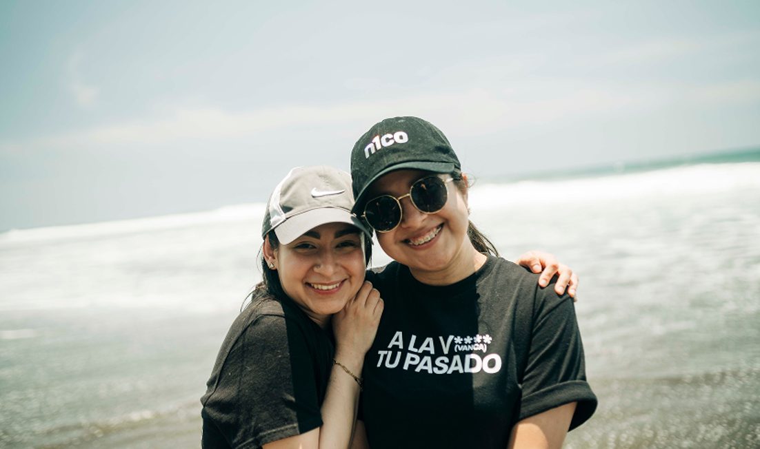 Dos jóvenes abrazadas sonríen en la playa, una con gorra de n1co
